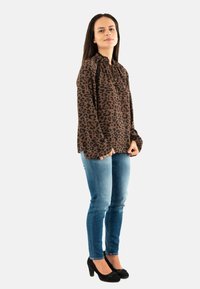 Donna che indossa una blusa marrone con stampa leopardata, jeans skinny blu e tacchi alti neri, in piedi con le mani unite davanti.