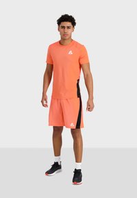 Pánský sportovní outfit v živé oranžové barvě; tričko s krátkým rukávem a šortky s černými postranními akcenty; vyrobeno z prodyšného materiálu.