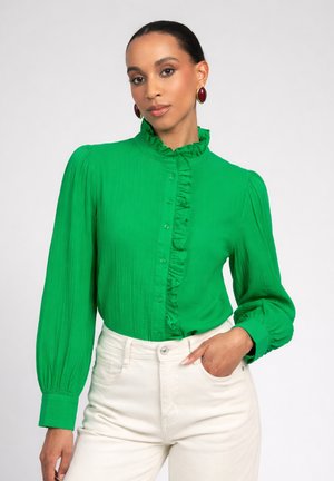 Femme portant un chemisier vert vif à volants avec des manches longues et un pantalon blanc taille haute, posant avec une main dans la poche.