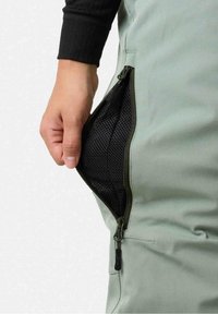 Main ouvrant la fermeture éclair latérale d’un pantalon vert clair révélant la doublure en filet noir de la poche, portant un haut noir à manches longues.