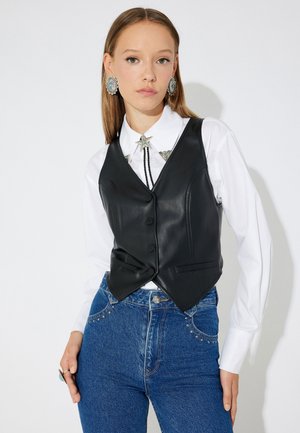 Gilet en cuir noir au design ajusté, avec un col en V, des boutons sur le devant et des poches à rabat. Porté sur une chemise blanche ornée.