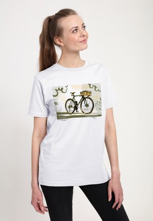 Kvinde iført en hvid T-shirt med et grafisk motiv af en sort cykel med en kurv mod en baggrund af abstrakte graffiti-stil mønstre.