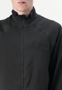 Juodas zip-up švarkas, pagamintas iš lengvos medžiagos, su aukštu gaubtu ir tekstūruotu logotipu kairėje krūtinės pusėje.