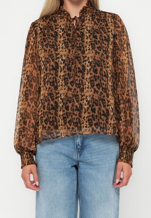 Frau, die eine braune und schwarze, langärmelige Bluse mit Leopardenmuster, Rüschenkragen und blaue, hoch taillierte Jeans trägt.