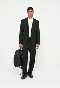Traje negro con chaqueta a medida y pantalones a juego, acompañado de una camisa blanca. Zapatos de cuero negros y una mochila negra.