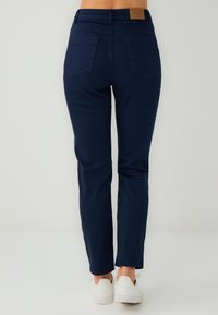 Persona che indossa jeans a gamba dritta blu scuro e sneakers bianche, in piedi contro uno sfondo chiaro e uniforme, visto da dietro.