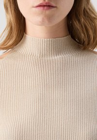 Beige bordázott kötött felső magas nyakkal és nyitott vállbetétekkel, sima textúrával és függőleges vonalakkal.
