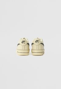 Os ténis Nike Air apresentam um acabamento superior leve em creme claro com detalhes pretos, superfície em pele texturizada, colarinho acolchoado e um design de sola plana clássico.