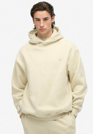 Jongeman met kort, donker haar, gekleed in een effen crèmekleurige hoodie en bijpassende broek, met handen in zijn zakken, tegen een witte achtergrond.