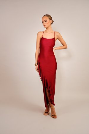 Femme portant une robe rouge foncé sans manches avec des volants et des sandales dorées à talons hauts, debout sur un fond uni.