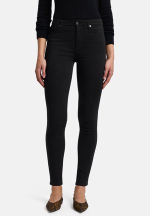 Femme portant un jean skinny noir taille haute, un haut noir à manches longues et des ballerines pointues imprimé léopard, debout devant un fond blanc uni.