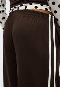 Κοντινό πλάνο καφέ παντελονιού με λευκές ρίγες στο πλάι, φορεμένο με μαύρο-άσπρο πουκάμισο με πουά, που δείχνει την ελαστική μέση και λεπτομέρειες τσέπης.