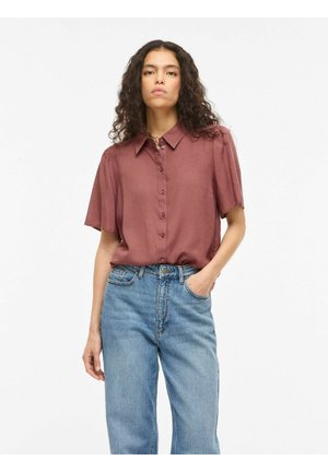 Femme aux cheveux bouclés portant un chemisier mauve à manches courtes boutonné, rentré dans un jean bleu taille haute, debout avec une main dans la poche.