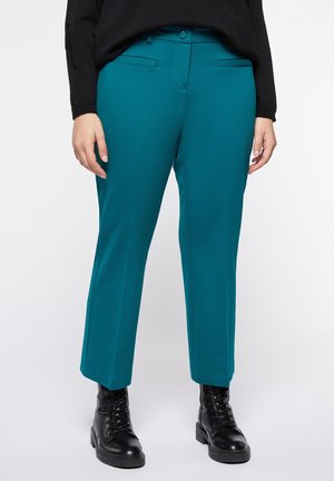 Pantalones entallados color azul verdoso hasta el tobillo, combinados con botas negras con cordones y un suéter negro, mostrados desde la cintura hasta los pies.