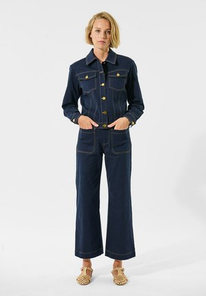 Veste en denim avec des boutons dorés, deux poches poitrine, assortie à un pantalon en denim évasé. Tissu bleu foncé avec des coutures jaunes contrastantes.