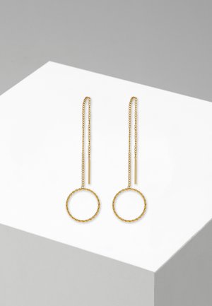 Gouden oorbellen met een gedraaide cirkelvormige ontwerp, voorzien van lange kettingen en cilindrische accenten, tentoongesteld op een witte geometrische voet.