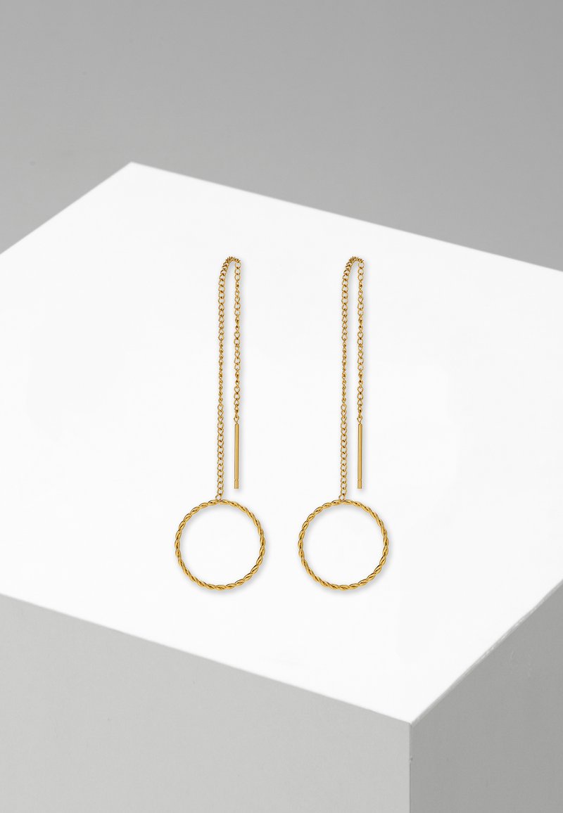 Orecchini d'oro con un design circolare intrecciato, caratterizzati da lunghe catene e accenti cilindrici, esposti su un piedistallo geometrico bianco.