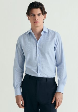 Joven con una camisa azul clara abotonada, metida en pantalones oscuros a medida, de pie frente a un fondo liso.