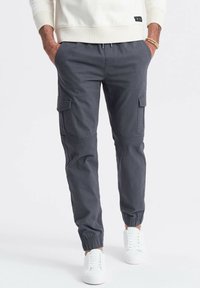 Grå cargo bukser med elastiske manchetter, flere sidelommer og en afslappet pasform, kombineret med hvide sneakers og en cremet sweatshirt.
