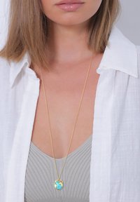 Gouden ketting met een ronde hanger waarop een blauw en gouden wereldbolontwerp is afgebeeld, gedragen over een grijs geribd topje, met een witte blouse.