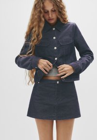Set de jachetă și fustă din denim de culoare bleumarin închis, dotat cu nasturi argintii, două buzunare pe piept și închidere cu nasture la fustă. Textură netedă.