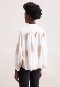 Blusa bianca con motivi floreali dorati, design fluente e schiena arricciata. Tessuto leggero con una vestibilità rilassata, caratterizzata da maniche lunghe.