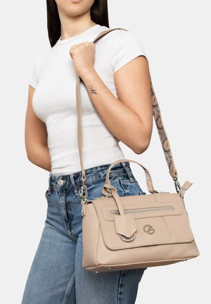 Frau in weißem Oberteil und blauer Jeans trägt eine beige Lederhandtasche mit gemustertem Schulterriemen und vorderer Reißverschlusstasche.