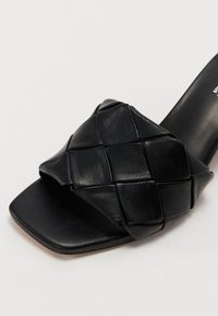 Black leather slide sandal featuring a woven strap design, square toe, and textured surface, with a smooth inner lining and subtle heel.