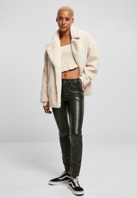 Getextureerde crèmekleurige bontjas, aansluitende lichtgroene crop top, glanzende zwarte leren broek en zwart-witte sneakers. Model staat met de handen langs het lichaam.