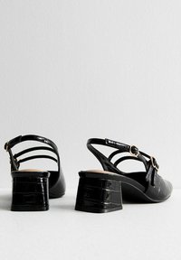 Svarta patentläder slingback-skor med blockklack och skurna remmar, med en rund spänndetalj och ett texturerat krokodilmönster.