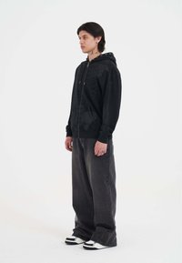 Sudadera negra con cremallera y estampado gráfico texturizado, combinada con pantalones oscuros de corte holgado en gris oscuro y zapatillas en blanco y negro.