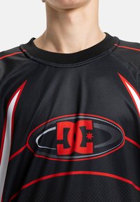 Melna sporta krekls ar sarkaniem un baltiem akcentiem, kas izceļ sarkano DC logo. Acs tekstūra un apaļš kakla izgriezums.