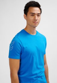 Blauwe katoenen t-shirt met korte mouwen. Beschikt over een ronde hals en drie chevronstrepen in wit op de linkerschouder. Soepele textuur.