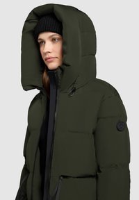 Padded parka bunda v tmavozelenej farbe s vysokým golierom, veľkou kapucňou, zipsovým uzatvorením a čiernou logom nášivkou na rukáve. Viditeľná čierna pletená čiapka.