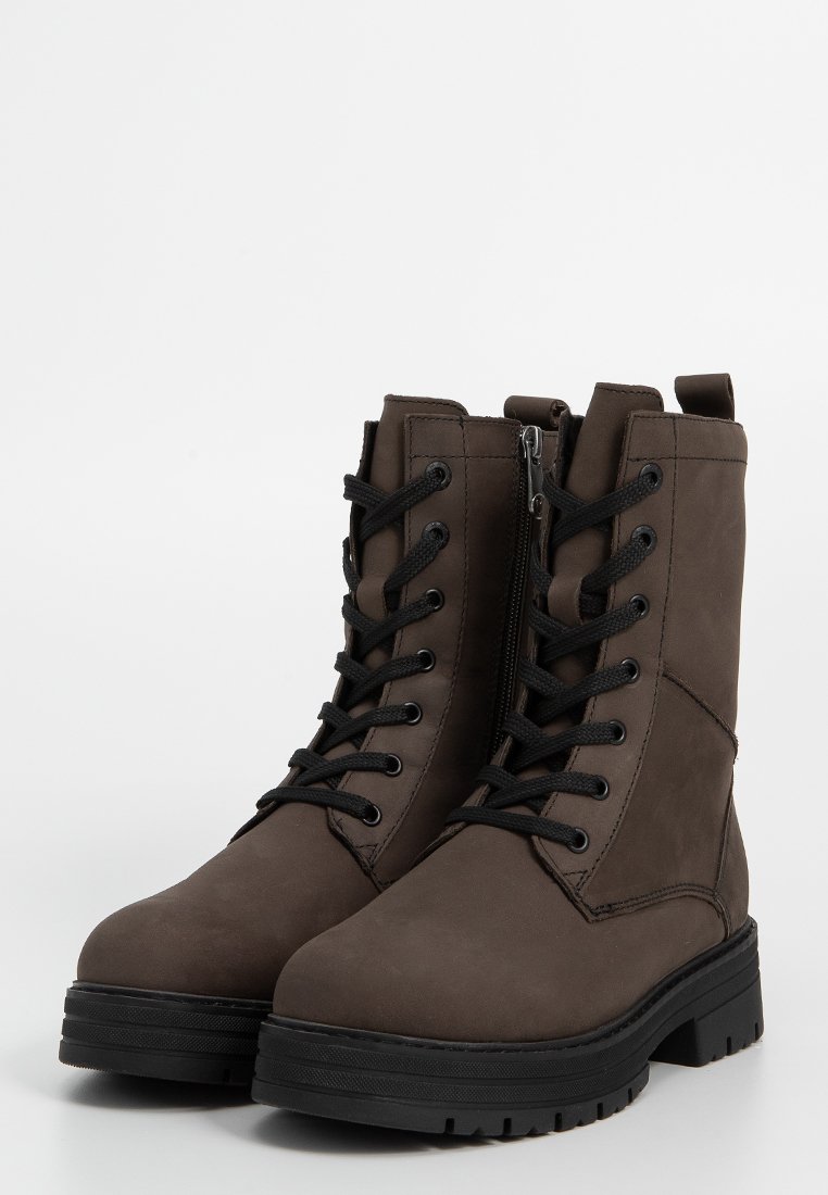 Par de botas de cuero marrón con cordones y suelas de goma negras gruesas, con cremallera lateral, de pie sobre una superficie blanca.