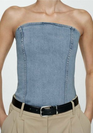 Osoba w błękitnym, jeansowym gorsetowym topie bez ramiączek, wpuszczonym w beżowe spodnie z czarnym paskiem i srebrną klamrą, z rękami w kieszeniach.