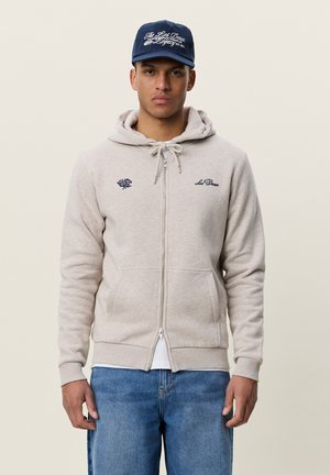 Jeune homme portant un sweat à capuche beige zippé avec des logos brodés, un jean bleu et une casquette bleu marine avec un texte manuscrit blanc.