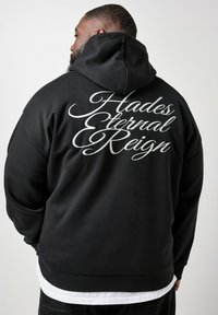 Hombre con sudadera negra con capucha y texto cursivo blanco "Hades Eternal Reign" en la espalda, de pie frente a un fondo claro y liso.