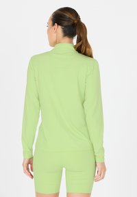 Femme avec queue de cheval brune portant un haut à manches longues vert clair et un short assorti, debout de dos à la caméra.