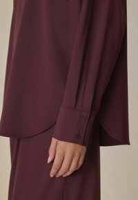 Burgundy long-sleeve blouse with a smooth texture, rounded hem, and buttoned cuffs. The design features a relaxed fit and flowing fabric.