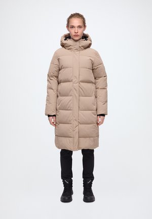 Une femme se tient face à l’objectif, vêtue d’un long manteau matelassé beige à capuche, d’un pantalon noir et de bottes noires, sur un fond blanc uni.
