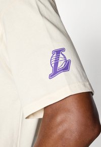 Kremfarget bomulls t-skjorte med en lilla brodert "L"-logo med basketball-design på venstre erme; kort ermer og avslappet passform.