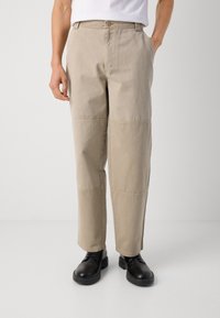 Beige cotton trousers with a relaxed fit, featuring side pockets and contrasting stitch detailing, paired with black shoes.