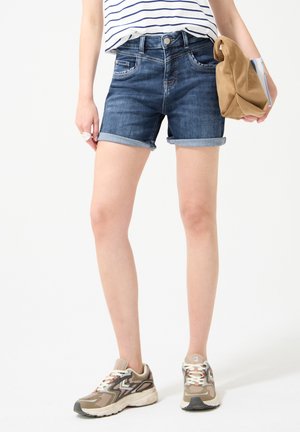 Personne portant un short en jean retroussé, un haut rayé, des baskets beige et marron, tenant un sac beige et un livre sur un fond blanc uni.
