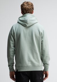 Mintgroene hoodie met een zachte textuur, voorzien van een grote capuchon en geribbelde manchetten, getoond vanaf de achterkant tegen een neutrale achtergrond.