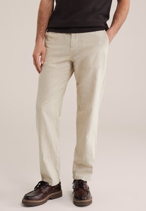 Bărbat purtând pantaloni bej, pantofi maro închis texturizați cu șireturi și cămașă neagră cu mâneci scurte, stând cu o mână în buzunar.