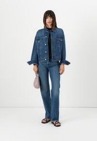 Veste en jean bleu foncé avec des boutons argentés, présentant des manches surdimensionnées et deux poches poitrine, associée à un jean fluide bleu clair et des sandales noires.