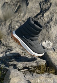 Cizmă de gleznă izolată, gri, cu un design matlasat, talpă din cauciuc, talpă intermediară albă și crampoane de culoare bej, așezată pe un teren rocky.