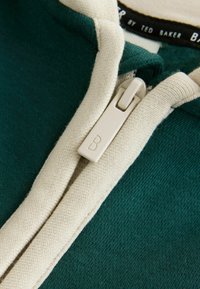 Sudadera verde con cremallera y ribetes color crema. Fabricada en una tela suave con un acabado texturizado. Presenta un tirador de cremallera marcado con una "B."