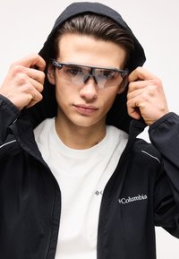 Jeune homme ajustant une veste Columbia noire à capuche, portant des lunettes de sport teintées et un t-shirt blanc sur fond uni.